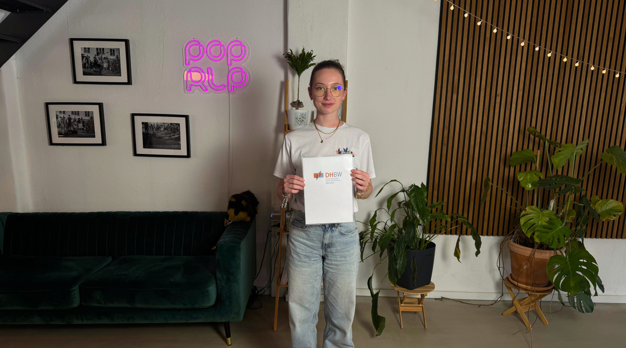 Emily steht im pop rlp Büro und hält Urkunde